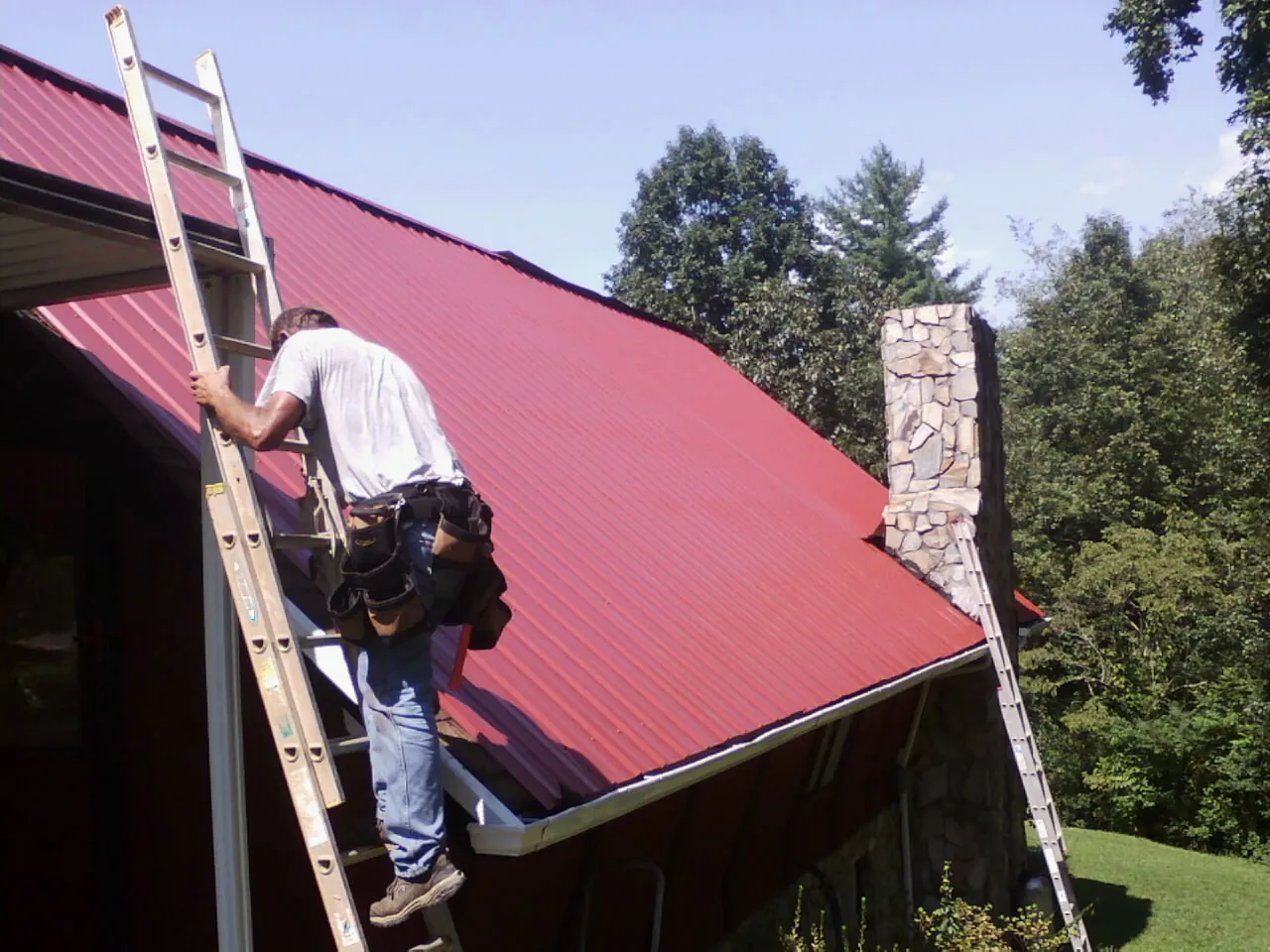 Roofer on a ladder beside a red metal roof and stone chimney — Soffit & Fascia Repair by Roofing Brothers in Albertson